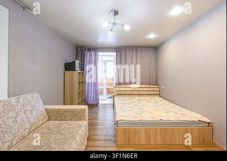 Gemütliches Zimmer mit einem gemusterten Sofa, einem Holzbett und helllila Vorhängen. Einfache Einrichtung, helle Beleuchtung. Stockfoto