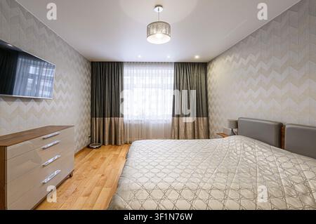 modernes Schlafzimmer zeigt ein Bett, eine Kommode und einen wandmontierten Fernseher. Der Raum ist in neutralen Tönen mit gemusterter Tapete und Holzböden dekoriert Stockfoto