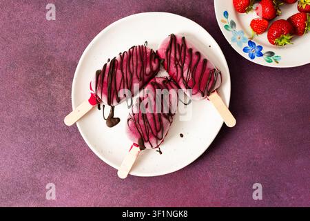 Drei Eis aus Erdbeeren, Bananen, Rüben und griechischem Joghurt sitzen auf einem Teller. Sie werden mit Schokolade getränkt und genießen während eines sonnigen Tages Stockfoto