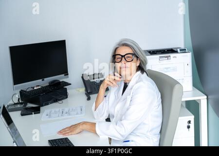 Erfahrene Ärztin lächelt im weißen Kittel und sitzt an ihrem Bürotisch Stockfoto