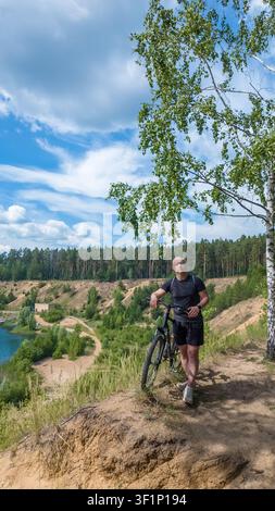 Mann steht neben einem Fahrrad in der Nähe eines Sees, der von Wald umgeben ist, an einem sonnigen Tag Stockfoto
