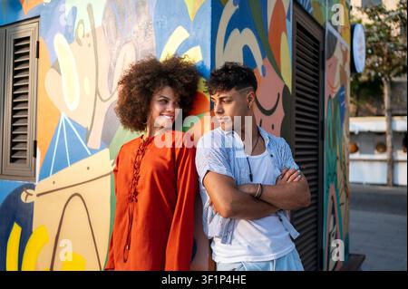 Stilvolles junges Paar in trendigen Sommeroutfits, das sich an einer farbenfrohen, künstlerischen Stadtmauer posiert Stockfoto