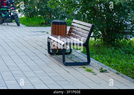 Eine ruhige städtische Szene mit einer Holzbank neben einem Mülleimer inmitten üppiger Vegetation auf einem gepflasterten Gehweg Stockfoto