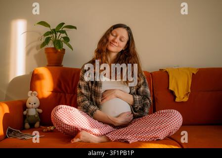 Schwangeren Mutter, die bequem auf einem Sofa sitzt, ihren Bauch liebevoll umschließt und Freude ausstrahlt, umgeben von Babyartikeln, während sie sich auf die Ankunft des Neuen vorbereitet und träumt Stockfoto