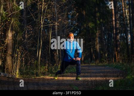 Mann in Sportkleidung führt einen tiefen Ausfallschritt auf einem sonnenbeschienenen Waldweg aus, konzentriert auf Dehnung und Aufwärmen vor einem Lauf, verkörpert Stärke, Ausdauer und Wohlbefinden Stockfoto