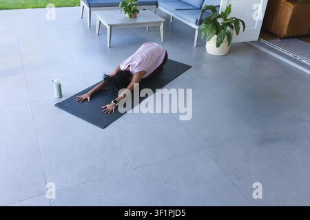 Afroamerikanische Frau kniet und dehnt sich auf einer dunklen Matte auf der Terrasse in einem rosa Oberteil, Leggings und einer Smartwatch Stockfoto