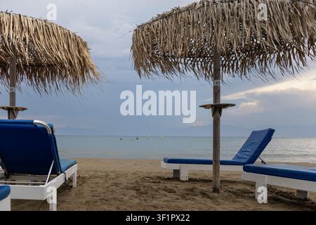 ruhige Strandszene mit nummerierten Strohschirmen und leeren Sonnenliegen unter einem stimmungsvollen, bewölkten Himmel über dem Meer Stockfoto