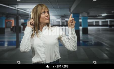 Mittelalter hispanische blonde Frau in weißem Hemd berührt ihr Ohr mit der linken Hand und zeigt den rechten Zeigefinger nach oben zwischen Betonsäulen und geparkten c Stockfoto