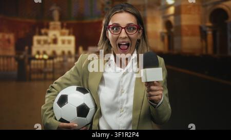 Reporterin hält Mikrofon und Fußball im Gebäude, Mund offen und spricht mit aufgeregter Pose in Richtung Kamera; Überraschungsfeier. Stockfoto