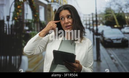 Mittlere ältere brünette Frau hält Tablet und zeigt mit dem Finger auf die Schläfe am Straßenrand; nachdenkliche Reflexion. Stockfoto