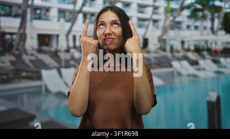 Die hispanische Frau zeigt mit beiden Zeigefingern nach oben auf die Liegestühle am Außenpool des Hotels, während sie lächelt; verspieltes Selbstvertrauen. Stockfoto