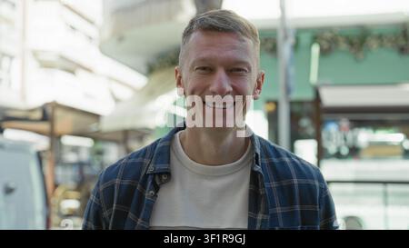 Mann in einem blauen karierten Hemd lächelt in die Kamera neben einem weißen Arbeitswagen mit Dachträger auf einer sonnendurchfluteten Fußgängerzone, die mit Cafés und Sonnenschirmen gesäumt ist; warm Stockfoto