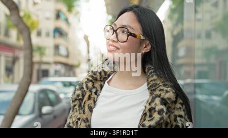 Frau mit Schildpatt-Brille und Leopardenmantel lächelt und blickt auf eine Stadtstraße in der Nähe geparkter Autos und Glasfassaden; Optimismus. Stockfoto