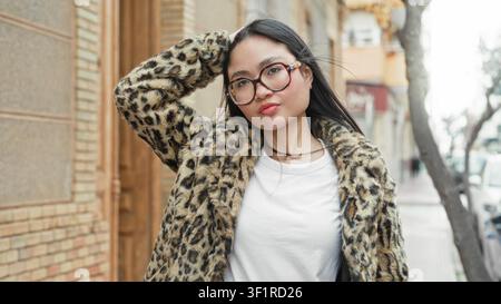 Frau mit Brille und Leopardenpelz lächelt mit der Hand hinter dem Kopf auf der Straße und lehnt sich an ein Gebäude; ungezwungenes Vertrauen. Stockfoto