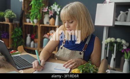 Blonde Floristin mit Schürze neben Laptop und Blumenstrauß in einem Floristikstudio; fokussierte Planung. Stockfoto