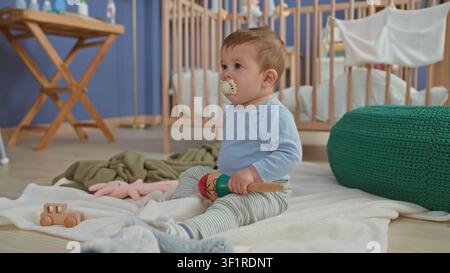 Junge, der auf einer Decke sitzt und eine hölzerne Maraca hält, Schnuller im Mund hält und ein Spielzeug mit der Hand hält, in einer Baumschule; sanfte Neugier. Stockfoto