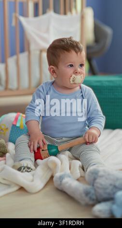 Kleinkind Junge, der auf einer Decke sitzt, Holzrassel hält und Plüschtier mit Schnuller im Mund in der Nähe eines Kinderbetts berührt; Neugier. Stockfoto