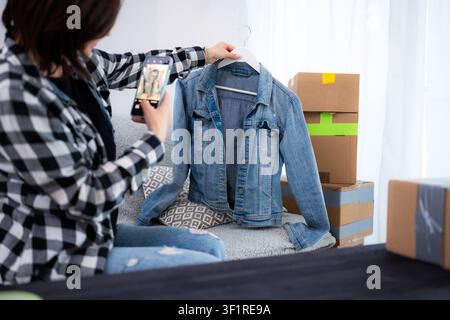 Die junge Frau macht Fotos Einer gebrauchten Jeansjacke für den Online-Verkauf und hält sie in der Hand Stockfoto