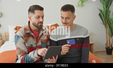 Der Mann hält das Tablet, während der Partner eine Kreditkarte hält und auf den Bildschirm auf einem Bett in einem hellen Schlafzimmer zeigt; Liebhabungsplanung. Stockfoto