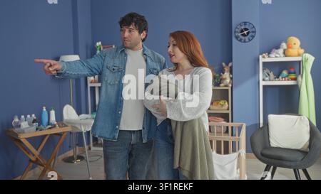 Frau, die ein in eine Decke gewickeltes Neugeborenes in einem blauen Kinderzimmer hält, mit einem lächelnden Mann neben einem Holzbett und einem Wickeltisch; Zärtlichkeit. Stockfoto