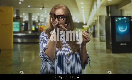 Eine Seniorin zeigt mit dem Finger auf eine Handbrille, während sie lächelt und ein Ersatzpaar in einem Flughafenterminal hält; Selbstvertrauen demonstriert. Stockfoto