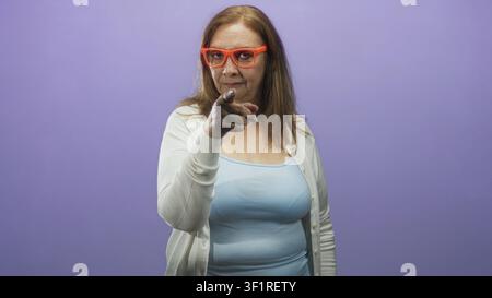 Frau zeigt mit dem Finger auf die Kamera in einem Studio, trägt eine orangefarbene Brille, eine weiße Strickjacke und ein blaues Tanktop mit direktem Blick; Autorität. Stockfoto