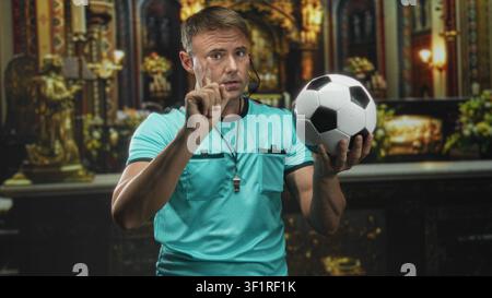 Mann Schiedsrichter hält Fußball und zeigt Finger, Pfeife auf Schlüsselband und Headset sichtbar in kunstvollen Gebäuden; Autoritätsdisziplin. Stockfoto