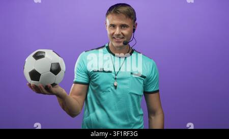 Der Schiedsrichter hält den Fußball in gestreckter Hand, mit Pfeife und Headset vor der violetten Studiowand; Selbstvertrauen. Stockfoto