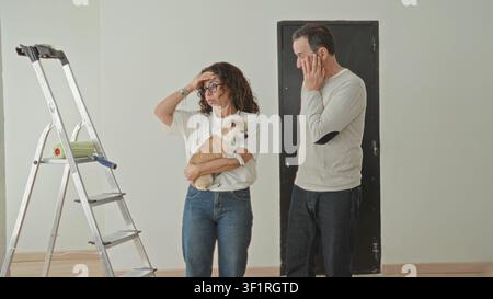 Frau, die einen kleinen Hund neben der Leiter hält, Hand an Stirn und Mann mit Hand an Hüfte im Gebäude; Sorge um Planungsentscheidung. Stockfoto