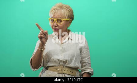 Frau mit gelber Brille berührt ihre Brust, hebt sich und zeigt mit dem Zeigefinger vor einem türkisfarbenen Hintergrund im Studio auf die Kamera Stockfoto