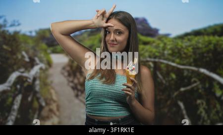 Die junge brünette Frau, die Eisbecher hält, lässt mich auf der Stirn ein Zeichen setzen, während sie auf einem Waldweg in der Nähe eines hölzernen Geländes steht; verspielter Sommerspaß. Stockfoto