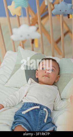 Hispanischer Junge, der im Kinderbett auf dem Bett liegt, Kopf auf gemustertem Kissen, trägt Jeansshorts und blickt auf das Plüschmobil, das über Holzlatten hängt Stockfoto