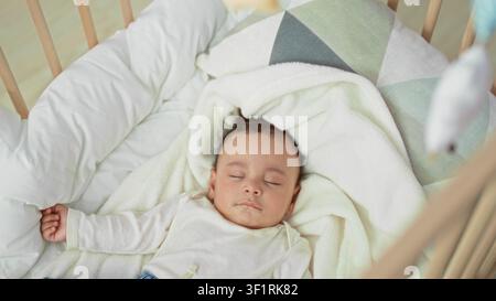 Baby schläft mit ausgestrecktem Arm und entspannter Hand in der Wiege im Kinderzimmer auf weißer Decke und geometrischem Kissen, Holzgeländer sichtbar; friedliche Ruhe. Stockfoto