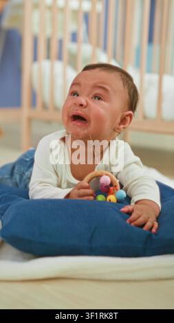 Hispanisches Baby, das eine Holzrassel hält, weint mit Tränen und Speichel, während der Betreuer sein Gesicht in einem Gebäude abwischt; Notfallbetreuung. Stockfoto