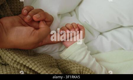 Kleiner Junge, dessen Hand den Finger eines Erwachsenen auf dem Bett umschließt, weißes Kissen und gestrickte Decke sichtbar; Bindungstendernheit. Stockfoto