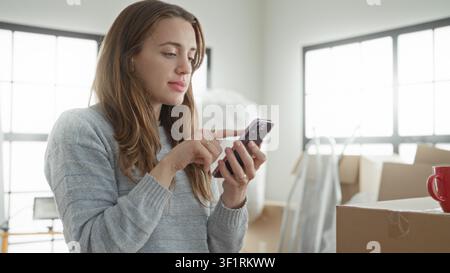 Frau, die im Studio neben gestapelten Kartonschachteln und Verpackungsmaterialien mit dem Finger auf das Smartphone klopft; ruhige Planung. Stockfoto