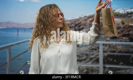 Frau in weißer Bluse und roter Brille hält kleine braune Geschenktüte aus Papier mit Herz von rechts oben auf dem Geländer am Seeufer; sanfte Nostalgie. Stockfoto