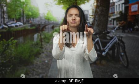 Frau zeigt beide Zeigefinger auf die Kamera auf der Kanalstraße in der Nähe eines geparkten Fahrrads in weißer Bluse; verspieltes Selbstvertrauen. Stockfoto
