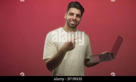 Lächelnder bärtiger Mann trägt gestreiftes T-Shirt, hält Laptop und zeigt mit dem Finger auf den Bildschirm im Studio; hilfreiches Online-Tutorial. Stockfoto