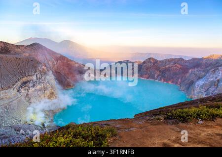 Panoramischer Blick auf den türkisfarbenen sauren Kratersee von Kawah Ijen mit aufsteigendem Schwefelrauch, umgeben von schroffen Vulkanfelsen bei Sonnenaufgang in Ost-Java, In Stockfoto