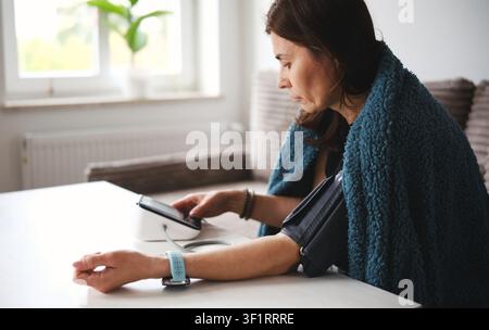 Frau, die auf einem Sofa in einer Decke sitzt, ein digitales Blutdruckmessgerät einschaltet und sich unwohl fühlt Stockfoto