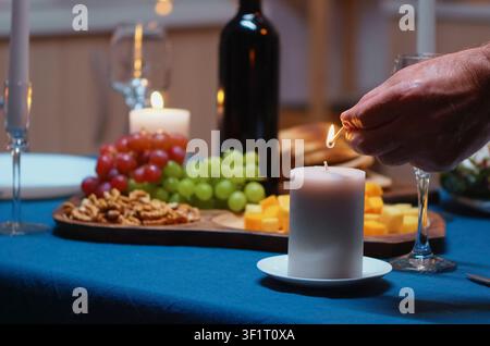 Ein älterer Mann zündet eine Kerze an, während er auf seine Frau bei einem romantischen Abendessen wartet, und bereitet ein festliches Essen für eine Jubiläumsfeier vor. Stockfoto