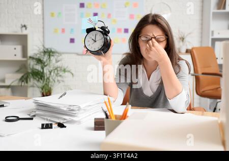 Gestresste junge Frau hält einen Wecker mit einem Fristzettel, erlebt Burnout und Druck in einer Büroumgebung Stockfoto
