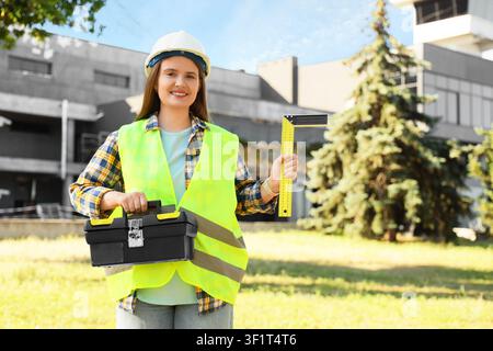 Lächelnde junge Ingenieurin mit Sicherheitshelm und Sicherheitsweste, die einen Werkzeugkasten und ein Lineal auf einer Baustelle hält, sie steht im Freien auf einer Baustelle Stockfoto