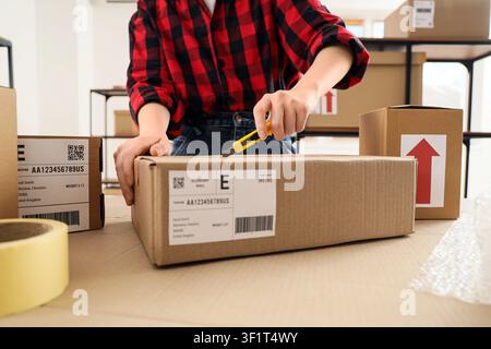 Person in kariertem Hemd öffnet einen Kartonversandkasten mit einem Universalmesser, E-Commerce- und Lieferdienstkonzept, der Kasten verfügt über ein Versandetikett Stockfoto