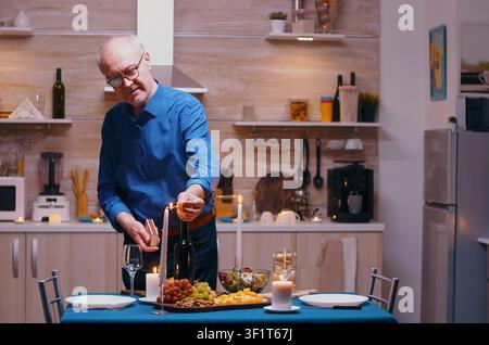 Ein Senior zündet eine Kerze an, während er auf seine Frau bei einem romantischen Abendessen wartet, und bereitet in der Küche ein festliches Jubiläumsessen mit gesunder Nahrung vor. Stockfoto
