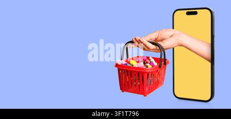 Hand, die einen roten Einkaufskorb voller farbenfroher Gegenstände hält, der aus einem Smartphone-Bildschirm herausragt, Online-Einzelhandel und E-Commerce Stockfoto