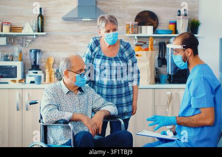 Arzt verwendet ein Infrarot-Thermometer, um einen behinderten Senior Mann im Rollstuhl während eines Hausbesuchs zur Gesundheitsversorgung zu überprüfen, während eines persönlichen Hauspflegebesuchs. Stockfoto