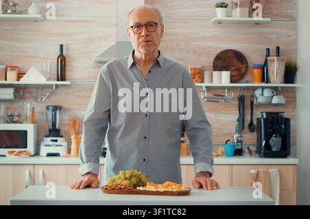 Senior Mann führt in der Küche ein Videoanruf, während die Familie hinter ihm das Abendessen zubereitet und Webcam-Technologie für Online-Gespräche verwendet. Stockfoto