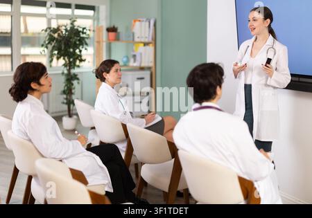 Selbstbewusste junge Frau Ärztin in weißem Mantel mit Medizinstudenten, die auf Stühlen sitzen, Ideen diskutieren und Präsentation mit zeigen Stockfoto